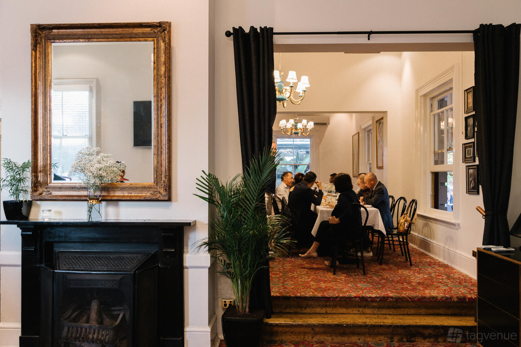 A dining room with chandeliers, large windows, and a fireplace with a framed mirror at The Mint Bar & Restaurant.