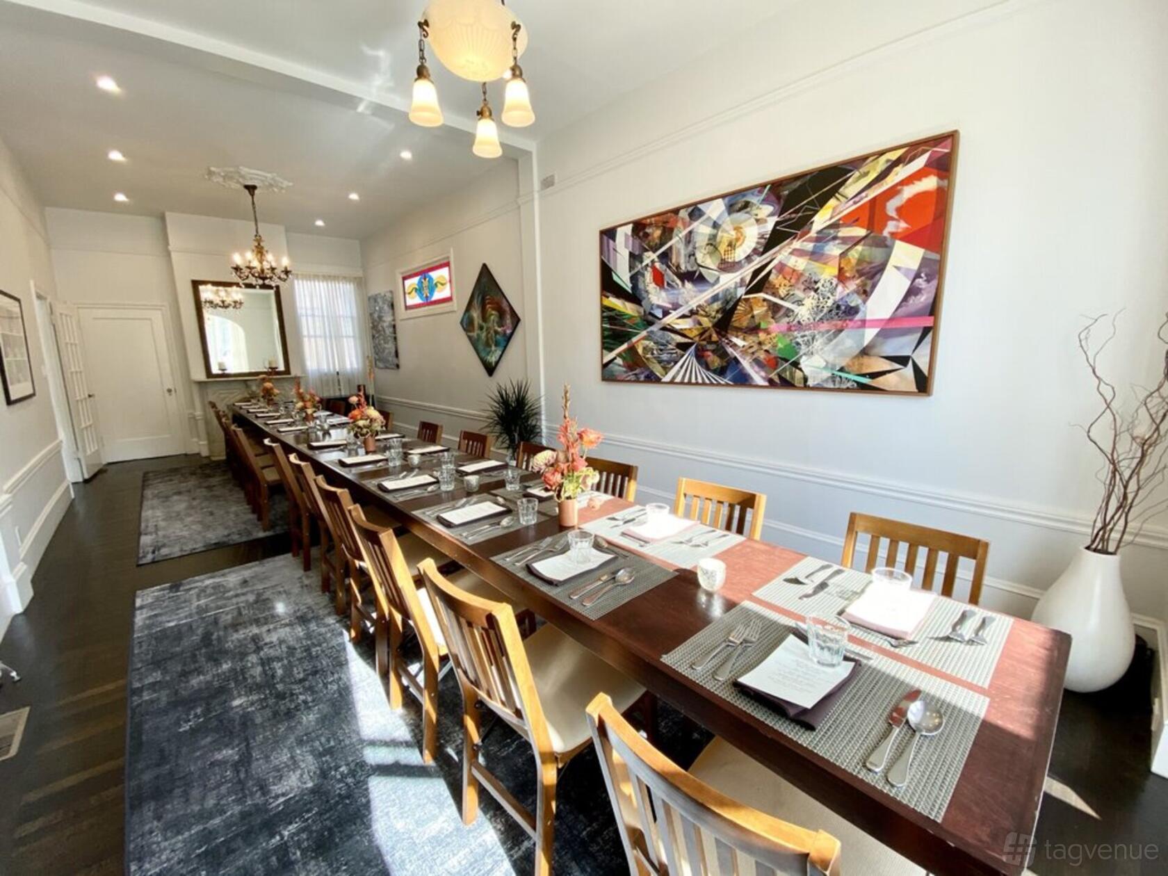 A private dining room with a long wooden table, wooden chairs, modern chandeliers, and colorful abstract art at Naked Kitchen.