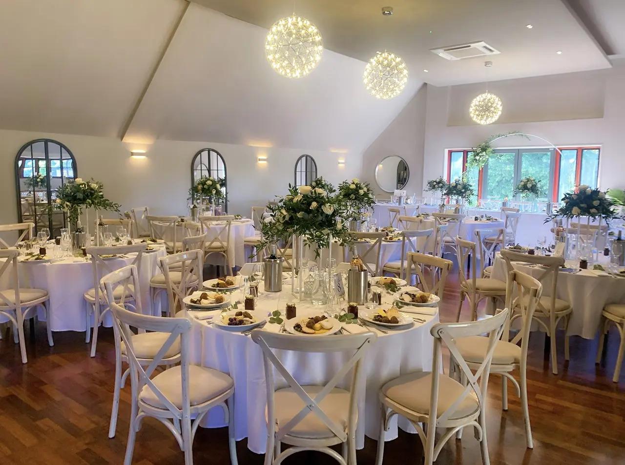 An event venue with round tables set for dining, white chairs, floral centerpieces, and globe pendant lighting at Hampton Court Palace Golf Club.