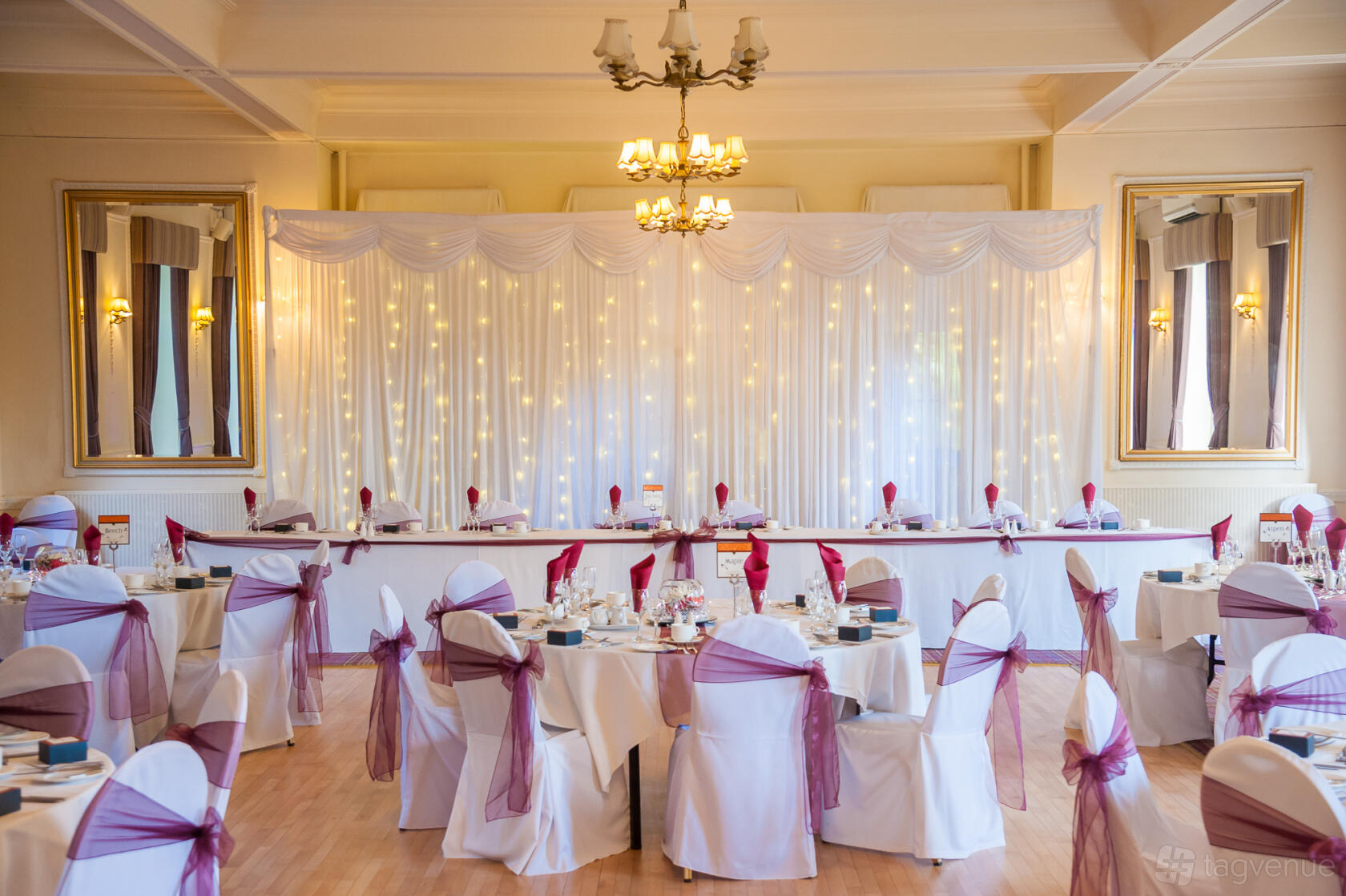 A country house dining room with round tables, white linens, mauve chair sashes, and a draped backdrop at Hoyle Court.