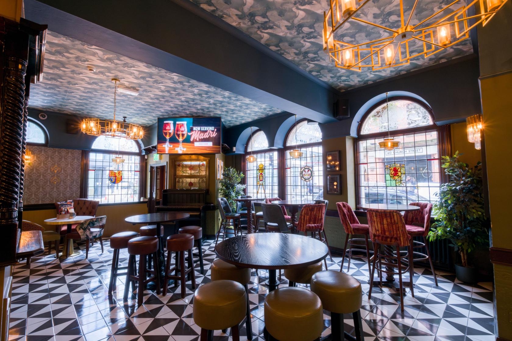A pub dining room with patterned tile floors, arched stained glass windows, and mixed seating at The Royal.