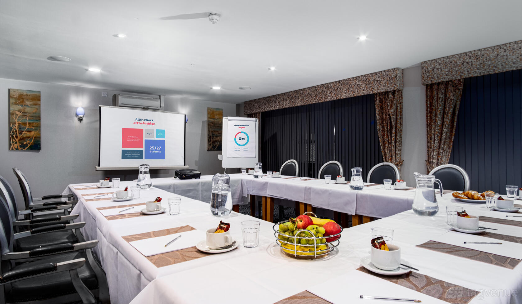 A meeting room with U-shaped tables, presentation screen, flip chart, and fruit basket at Cotswolds Hotel & Spa.