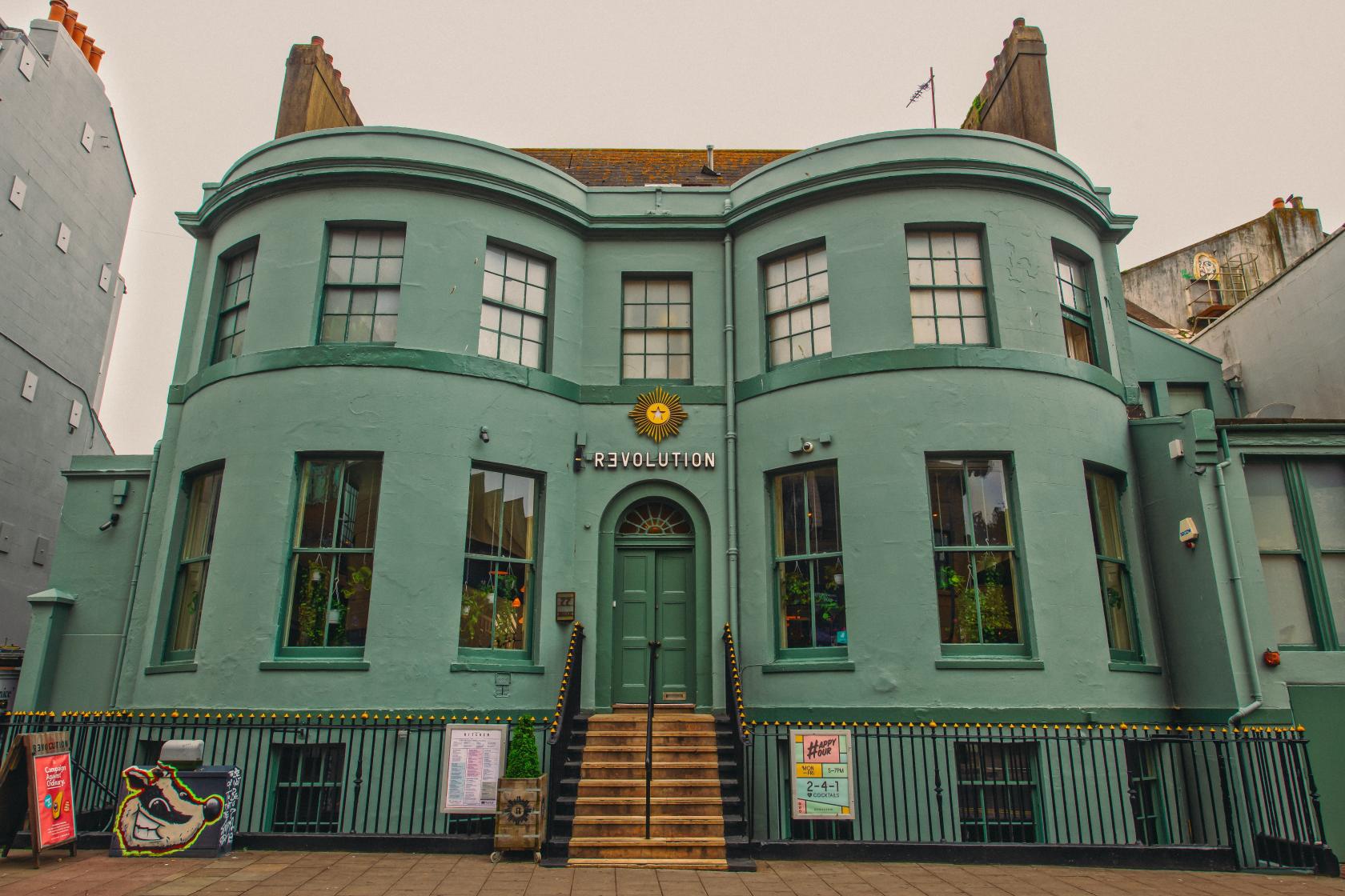 A green historic building with large arched windows, central steps, and art deco details at Revolution Brighton.