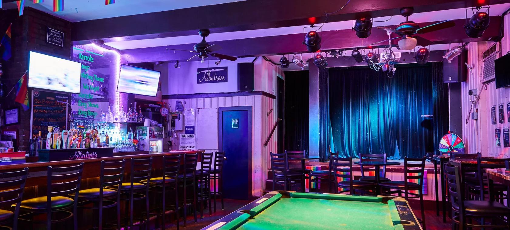An indoor bar with a stage, pool table, bar seating, neon lighting, and wall-mounted TV at Albatross Bar.