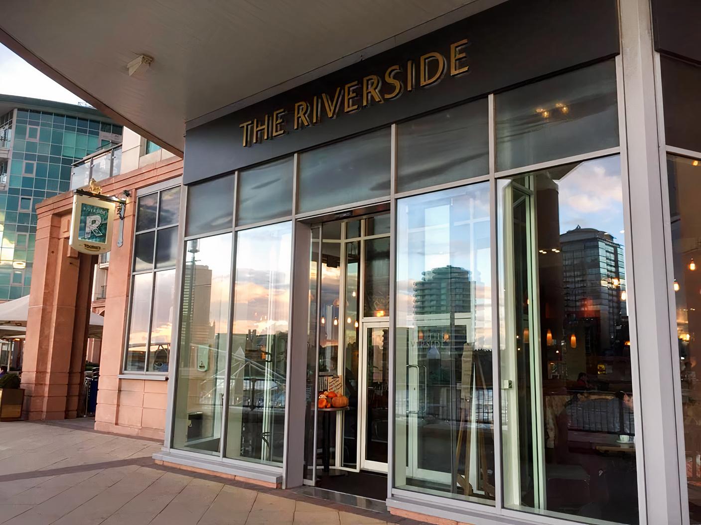 A restaurant entrance with floor-to-ceiling glass windows, modern signage, and city reflections at The Riverside.