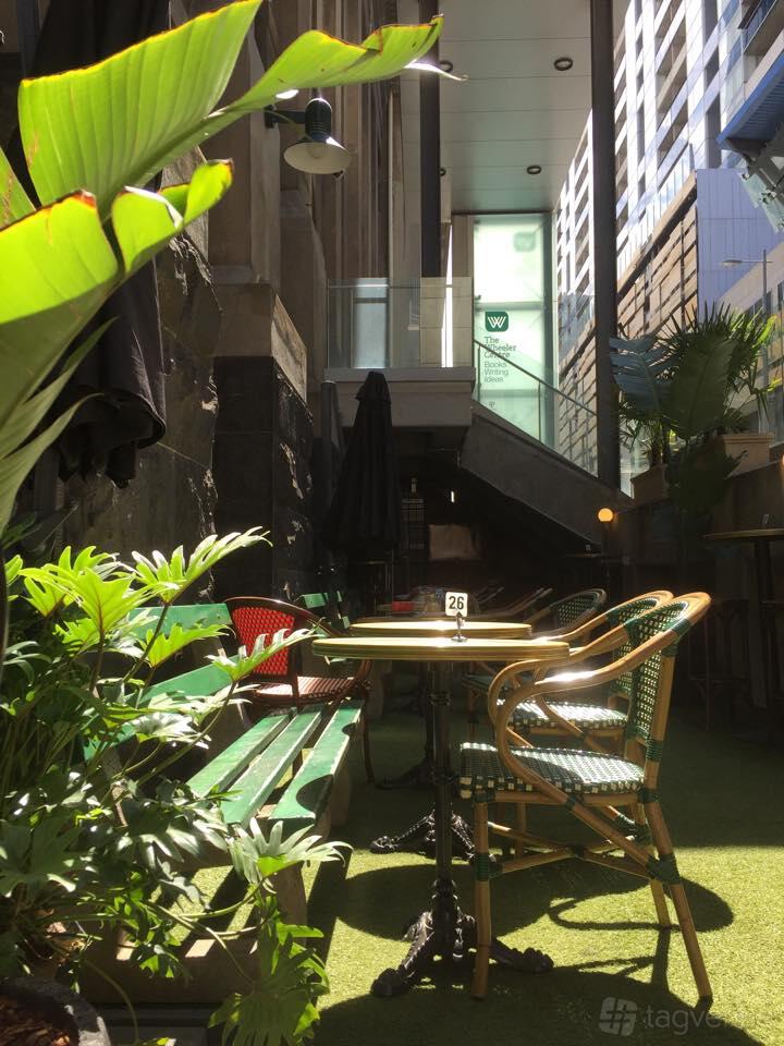 A restaurant patio with green wicker chairs, round tables, leafy plants, and brick walls at The Moat.