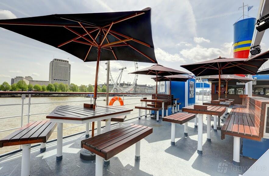 An open-air boat deck with wooden picnic tables, large black umbrellas, and riverside views at The Tattershall Castle.