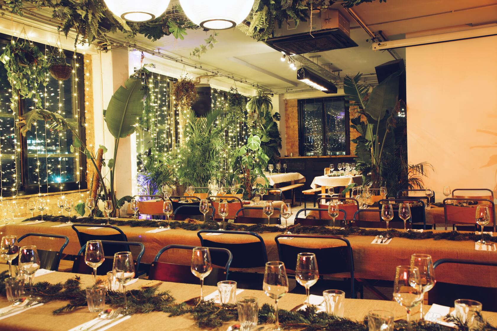 A cocktail bar with long wooden tables, fairy lights, potted plants, and hanging greenery at NT's Loft.