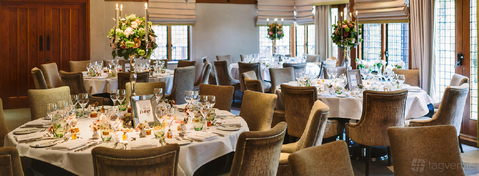 A hotel dining room with round tables, beige upholstered chairs, floral centerpieces, and large windows at Langshott Manor Hotel.