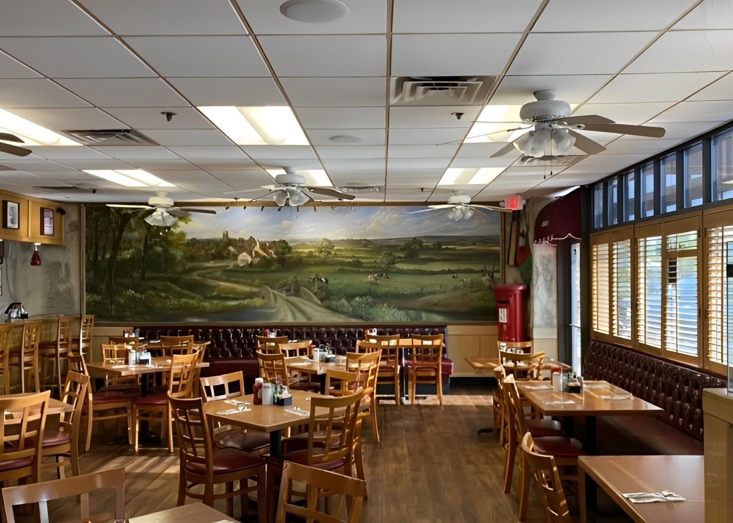A restaurant dining area with wooden tables and chairs, a countryside mural, and large windows at Annie's Cafe Lake Elsinore.