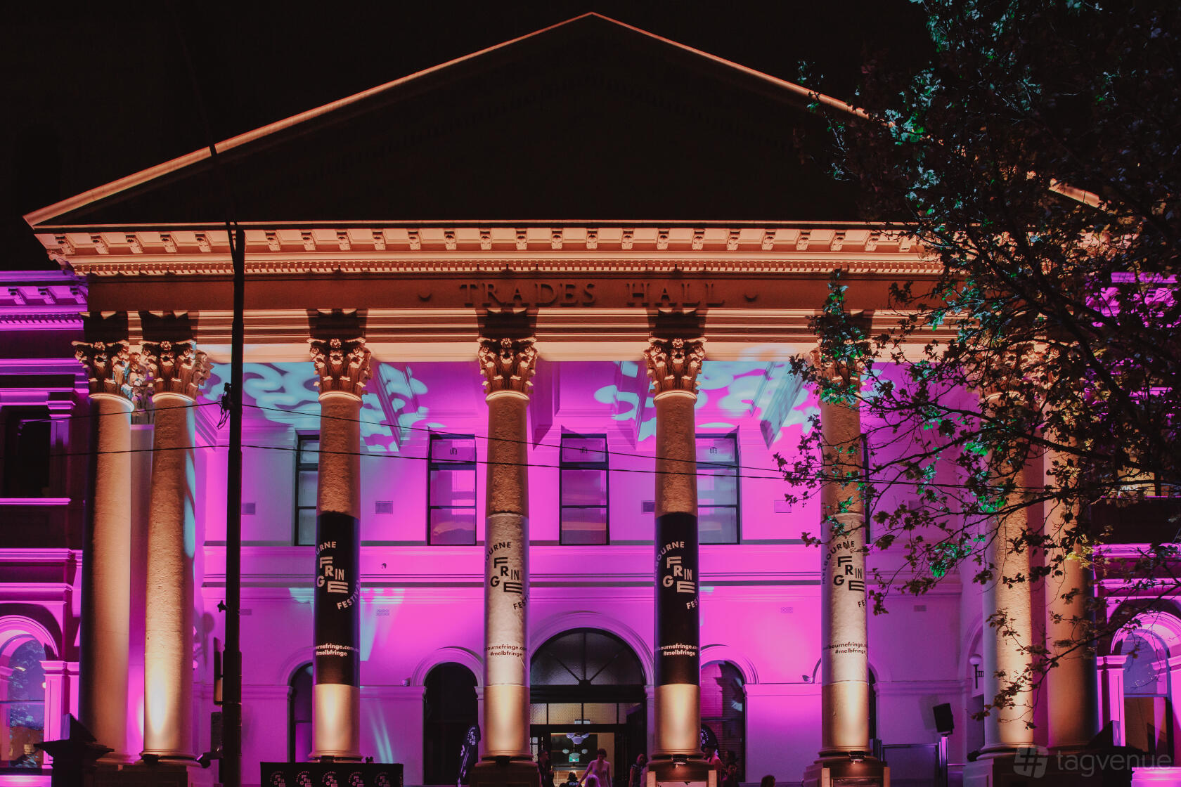 A neoclassical building facade with illuminated columns and colorful pink and purple lighting at Fringe Common Rooms.