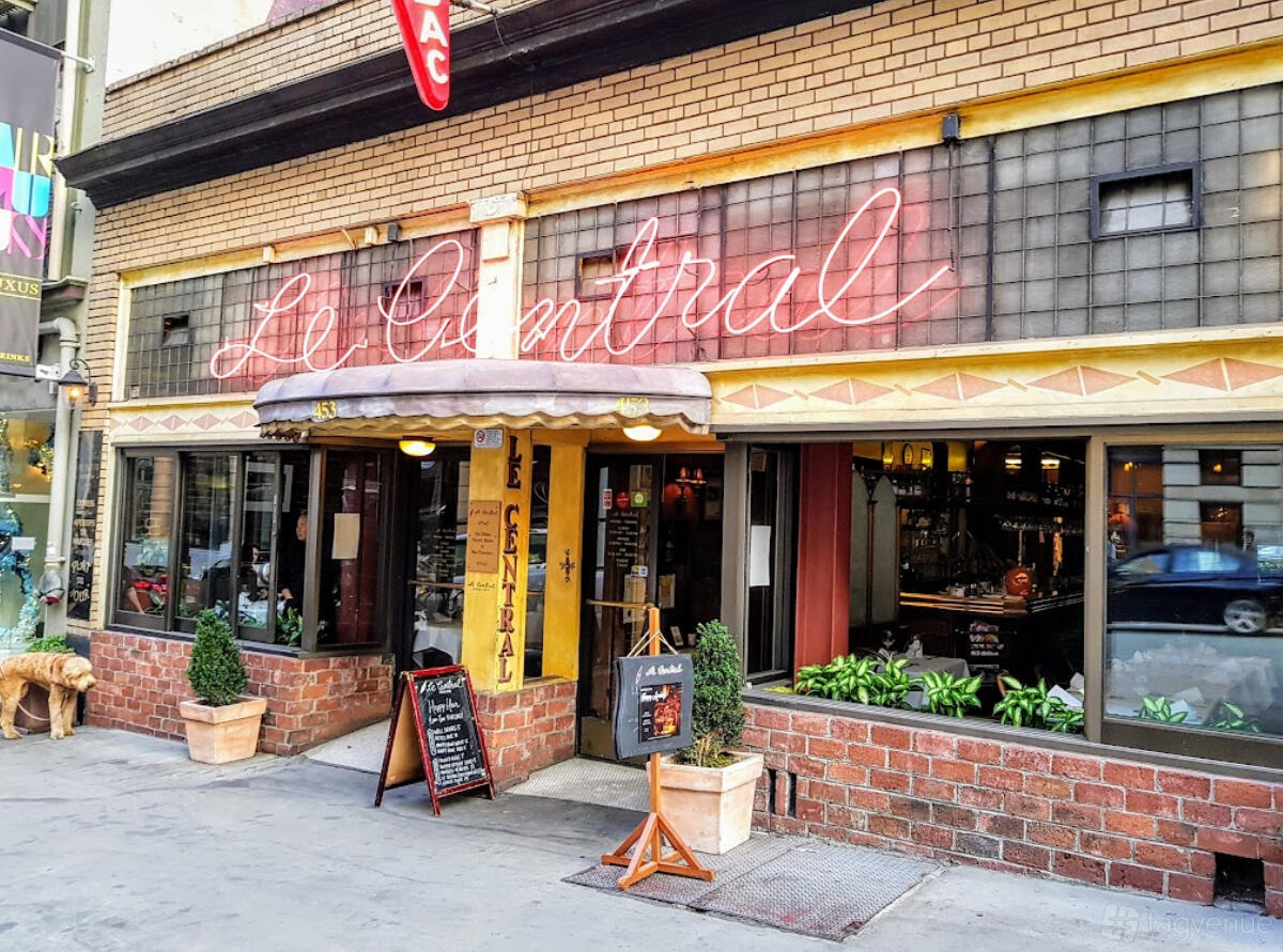 A restaurant with a neon sign, striped awning, large front windows, and potted plants at Le Central Bistro.