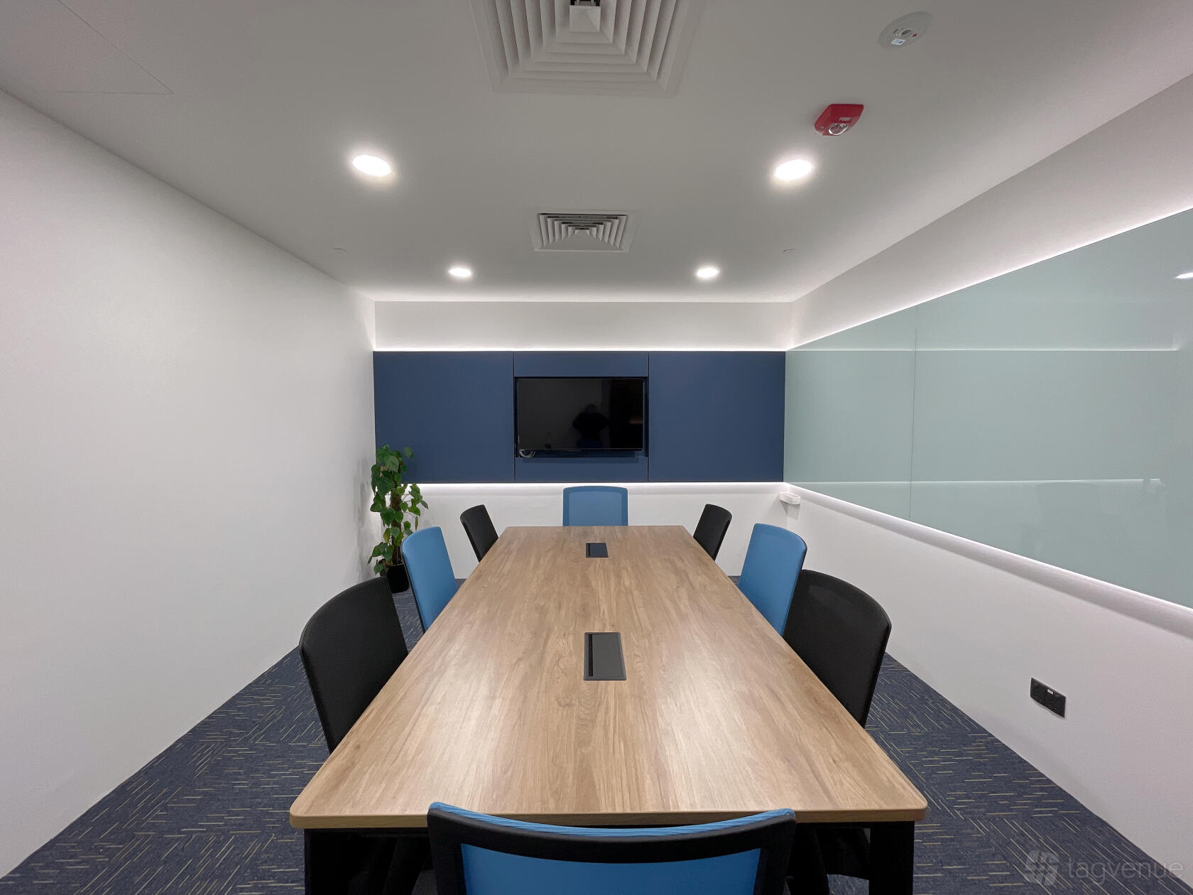 A meeting room with a long wooden table, black and blue chairs, wall-mounted TV, and a glass board at Le Space.