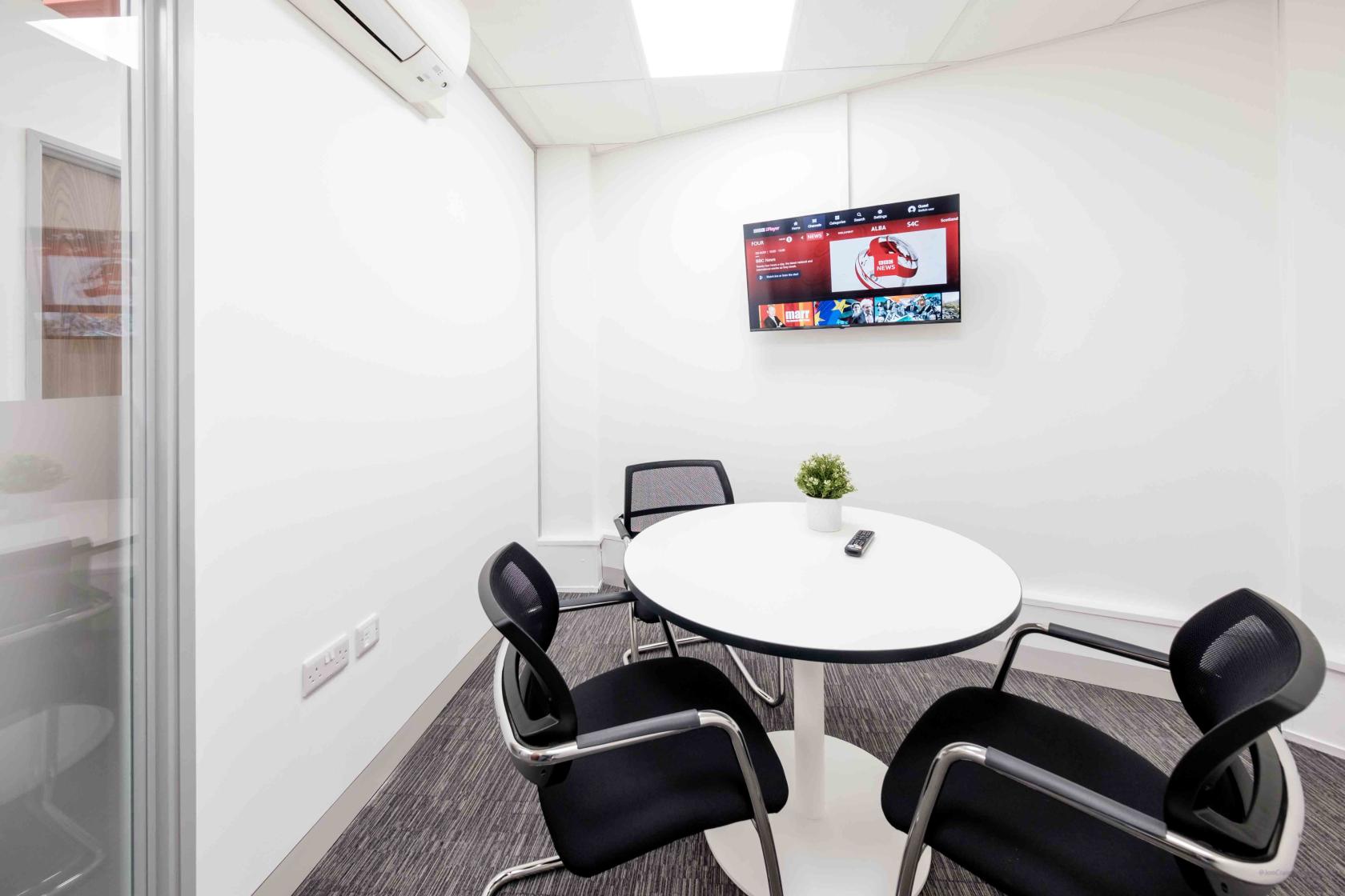 A small meeting room with a round white table, three black mesh chairs, and a wall-mounted display screen at Newminster House.