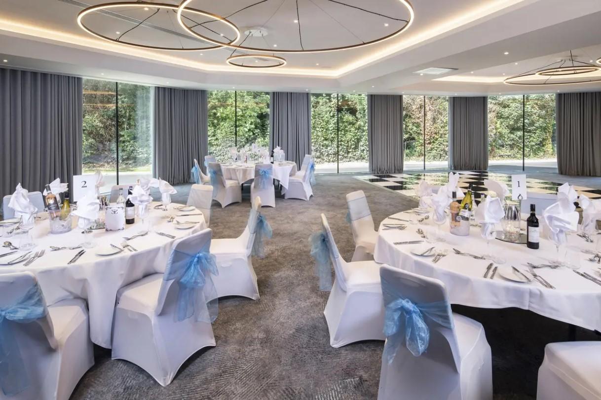 A meeting room with round tables covered in white linens, blue chair sashes, and floor-to-ceiling windows at Holiday Inn Shepperton.