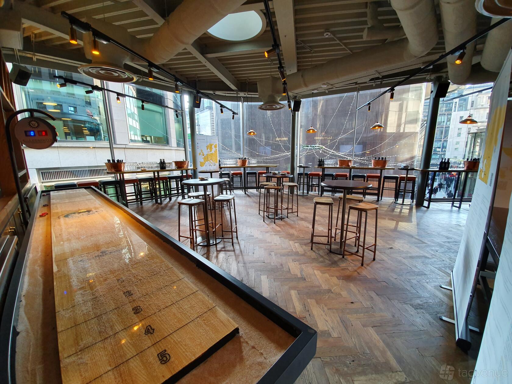 An event space in a pub with floor-to-ceiling windows, high tables, stools, and a shuffleboard table at Broadwood.