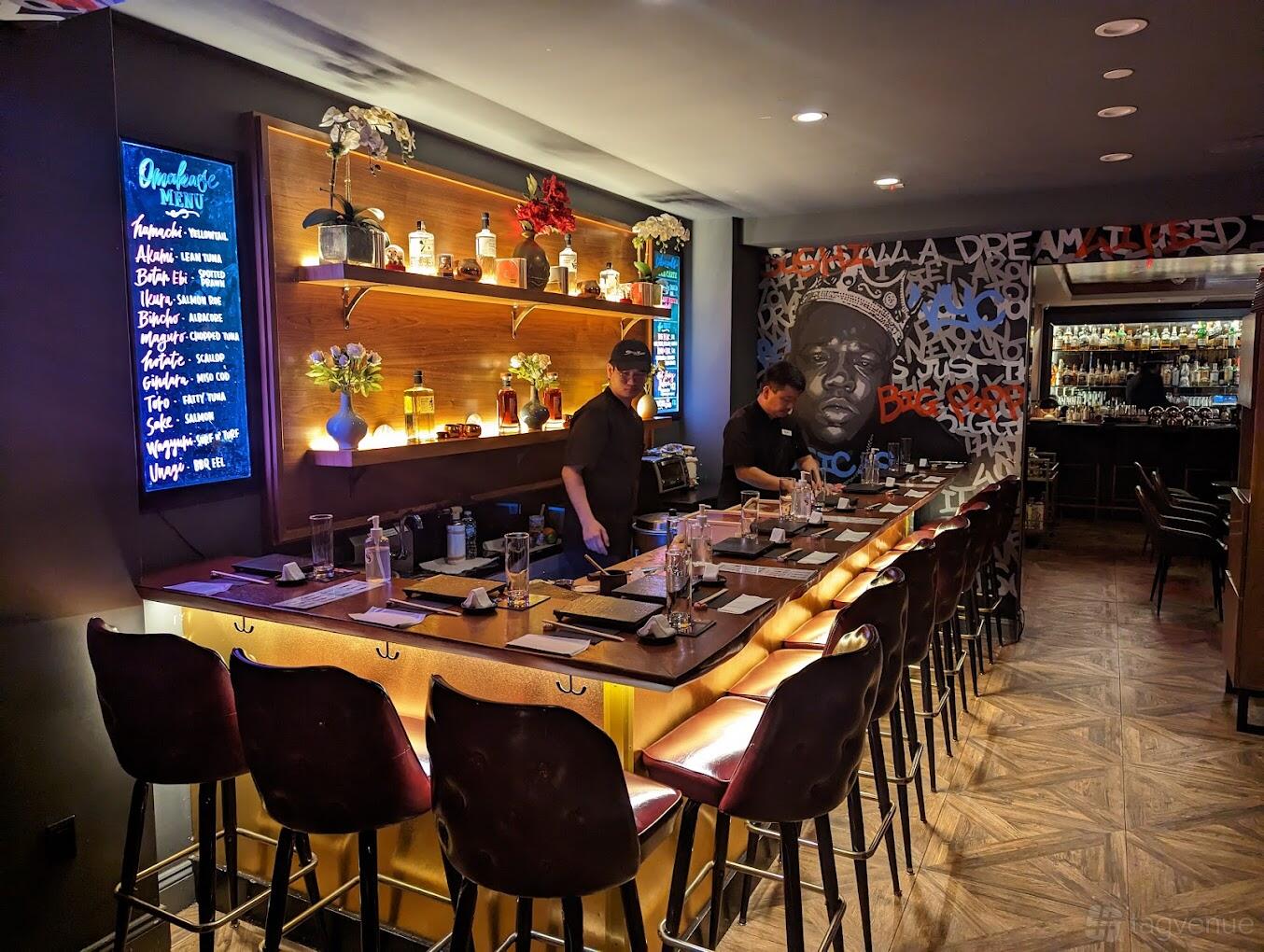 A restaurant with an illuminated sushi bar, wall shelves displaying bottles, and graffiti mural at Sushi by Bou Nomad.