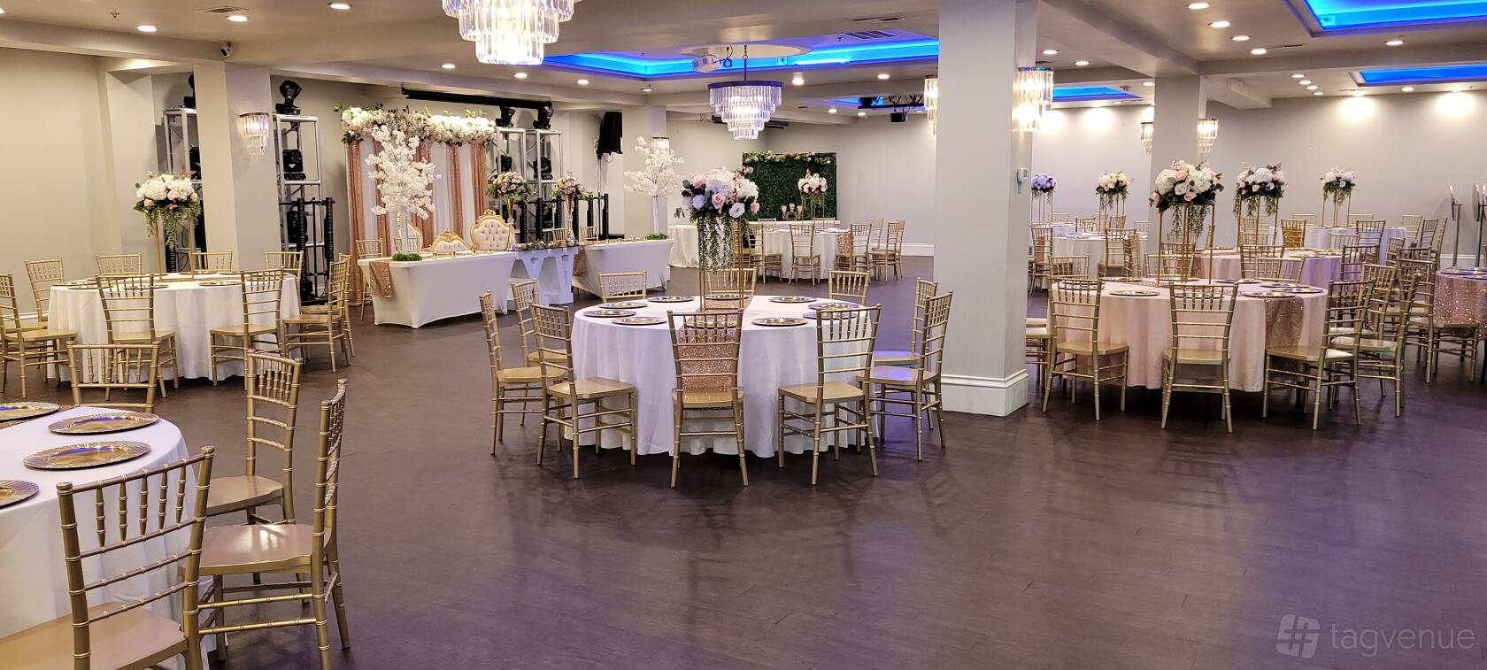 A hall with gold chiavari chairs, round tables with white linens, floral centerpieces, and chandeliers at Grand Luxe Hall.