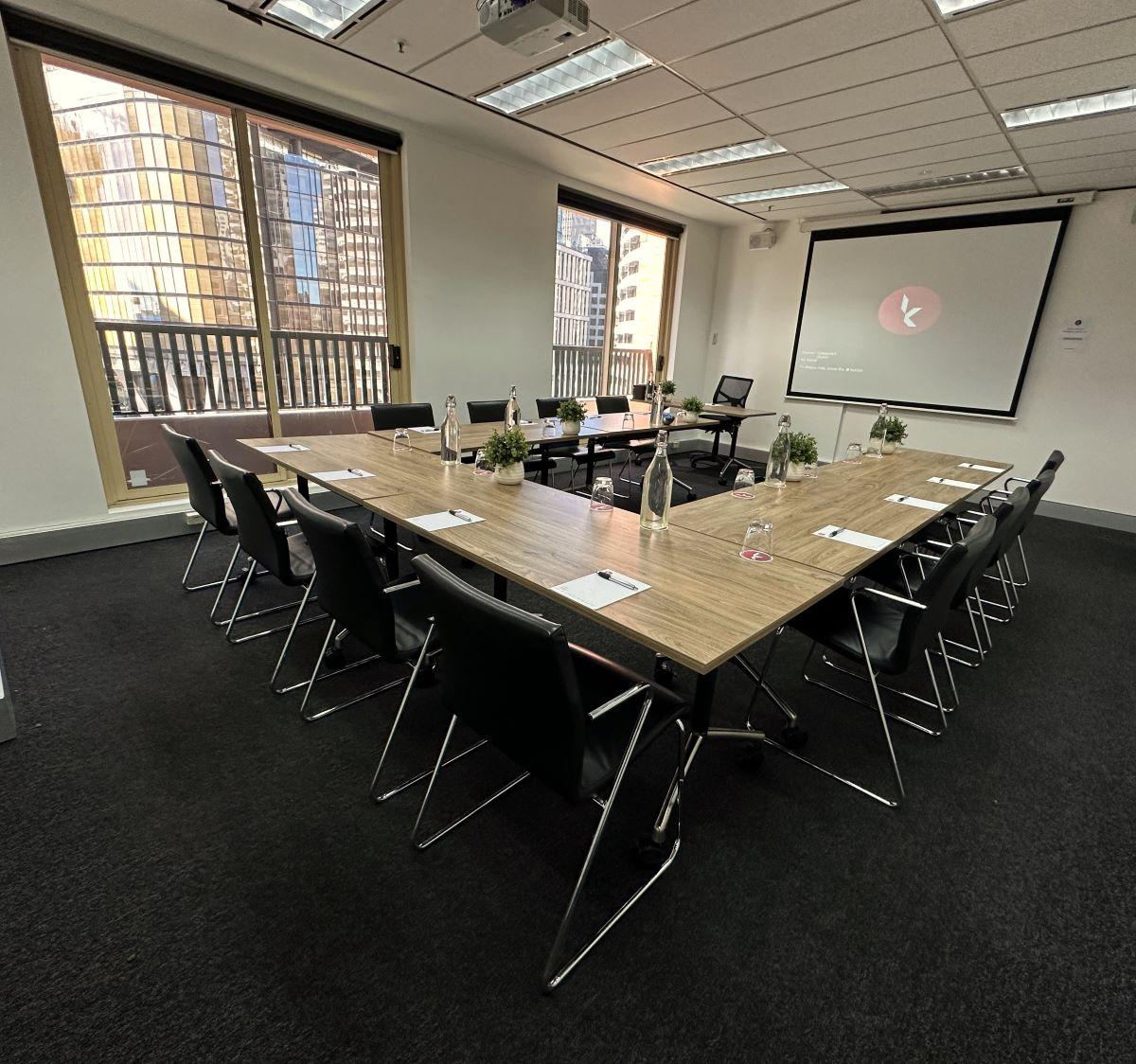 A conference centre boardroom with large windows, U-shaped tables, black chairs, and presentation screen at Karstens Sydney.