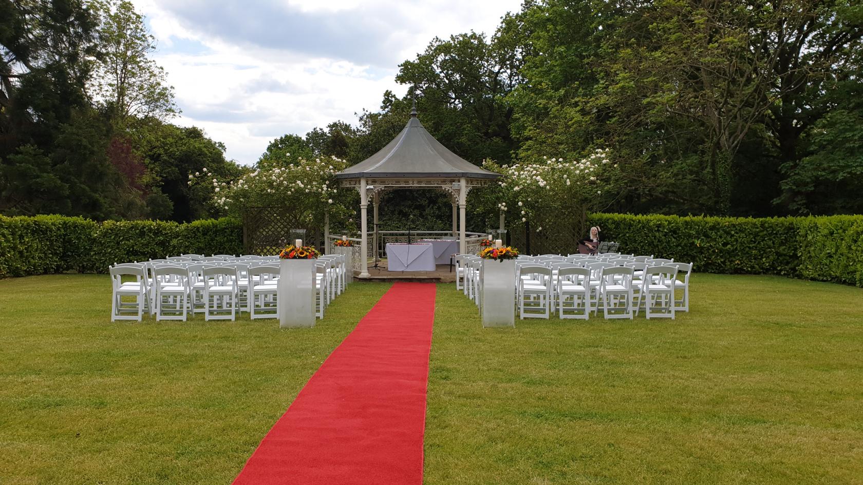 An outdoor event space with white chairs, a red carpet aisle, and a gazebo in the garden at Farnham House Hotel.