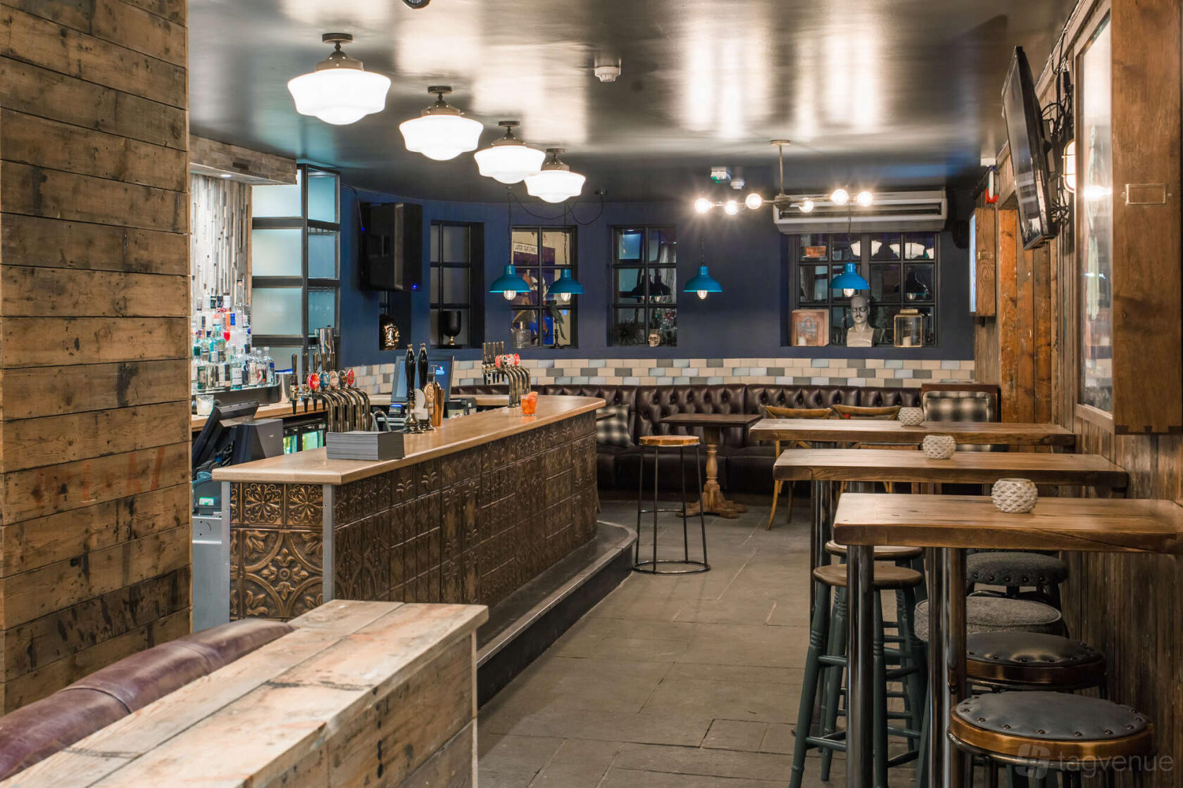 An event space in a pub with a central ornate bar, rustic wood paneling, leather banquettes, and pendant lighting at Duke's Head Putney.