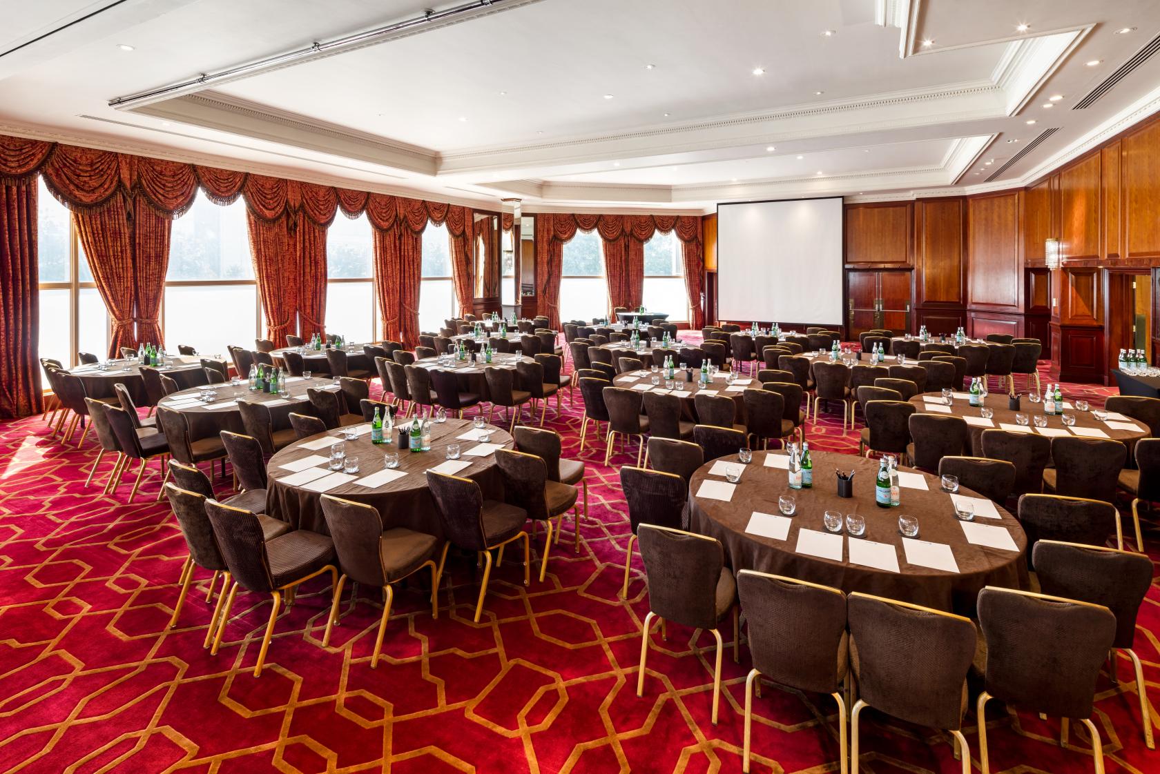 A function room with round tables, brown chairs, red carpet, and large windows with draped curtains at Radisson Blu Hotel, London Heathrow.