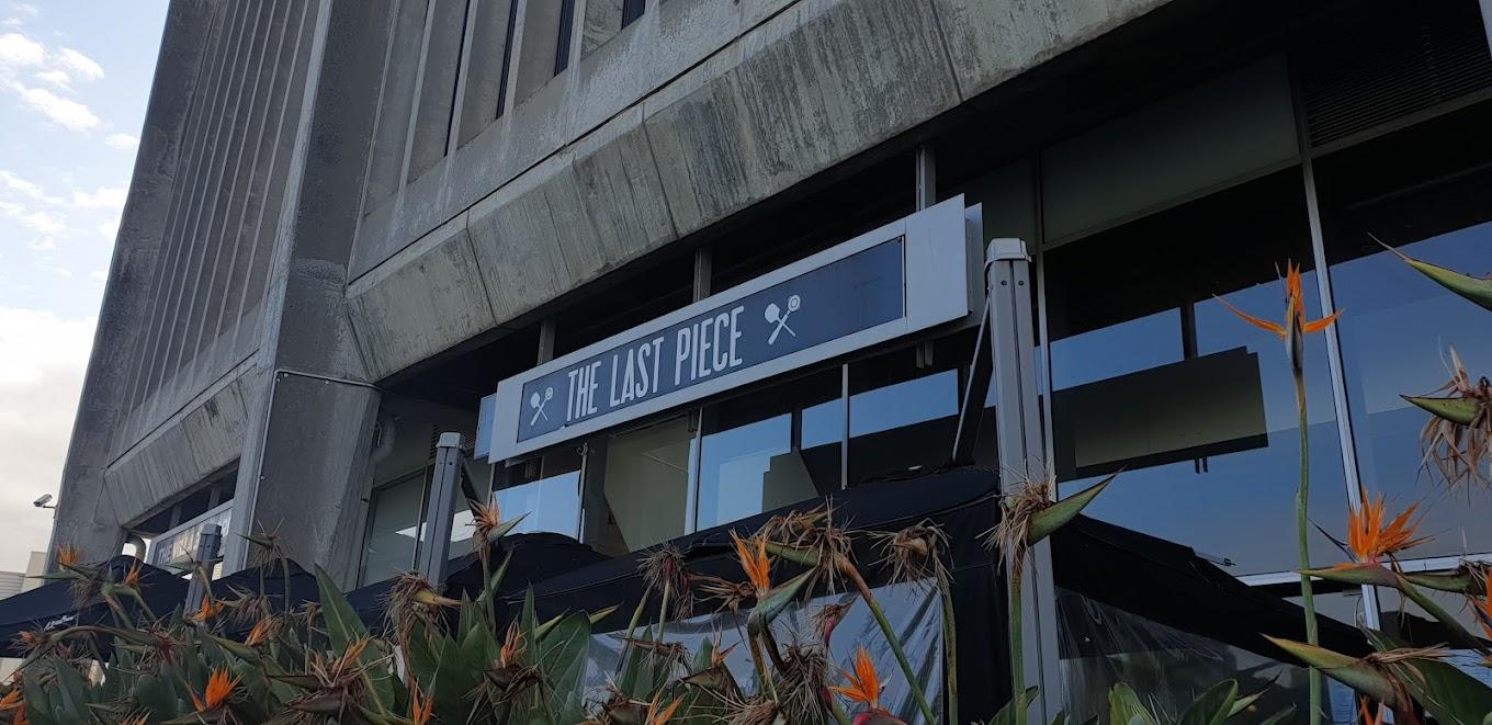 A restaurant exterior with concrete walls, large windows, and a sign reading 'The Last Piece' above the entrance at The Last Piece Restaurant.