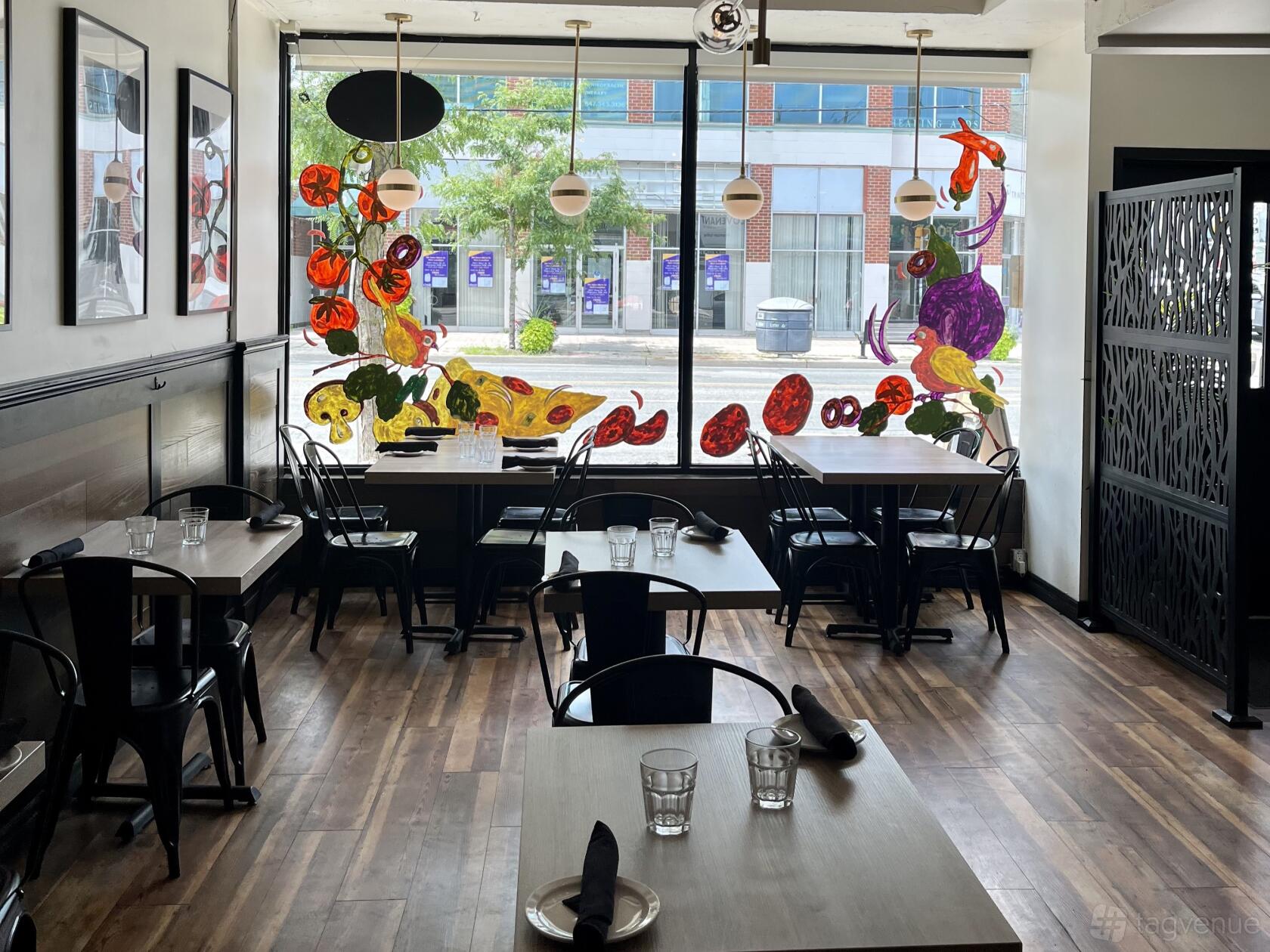 A restaurant dining area with wood floors, black chairs, set tables, and vegetable art on a large front window at Pizzeria Via Napoli.