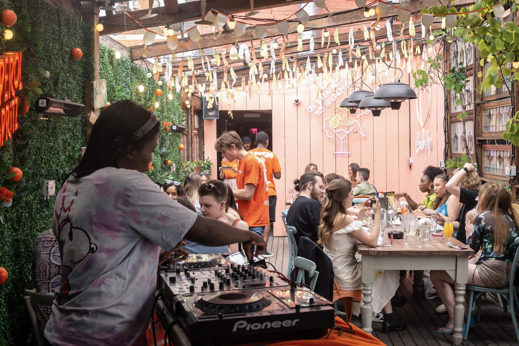 A rooftop bar with wooden decking, DJ booth, overhead string lights, and guests dining at Big Chill Kings Cross.