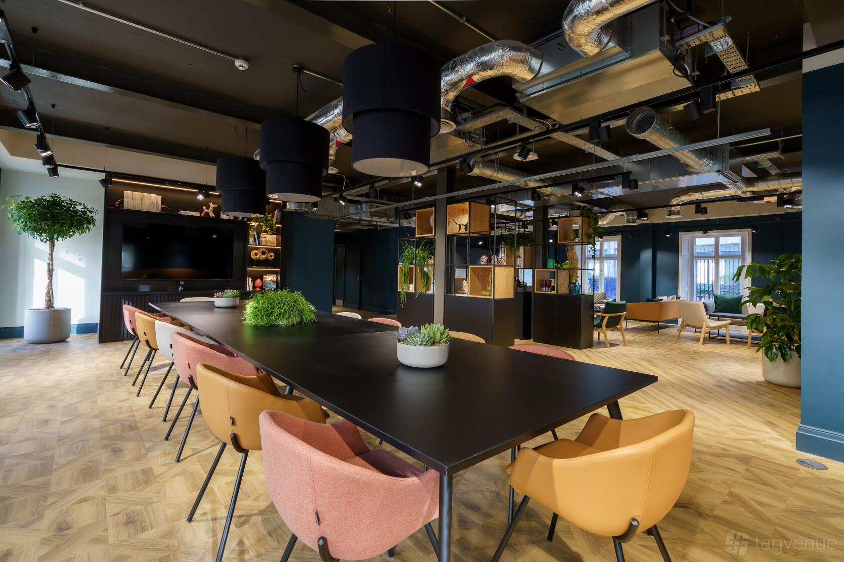 An event space with a long black table, pastel chairs, potted plants, and exposed ceiling ducts at Wizu Workspace - Newcastle - Portland House.