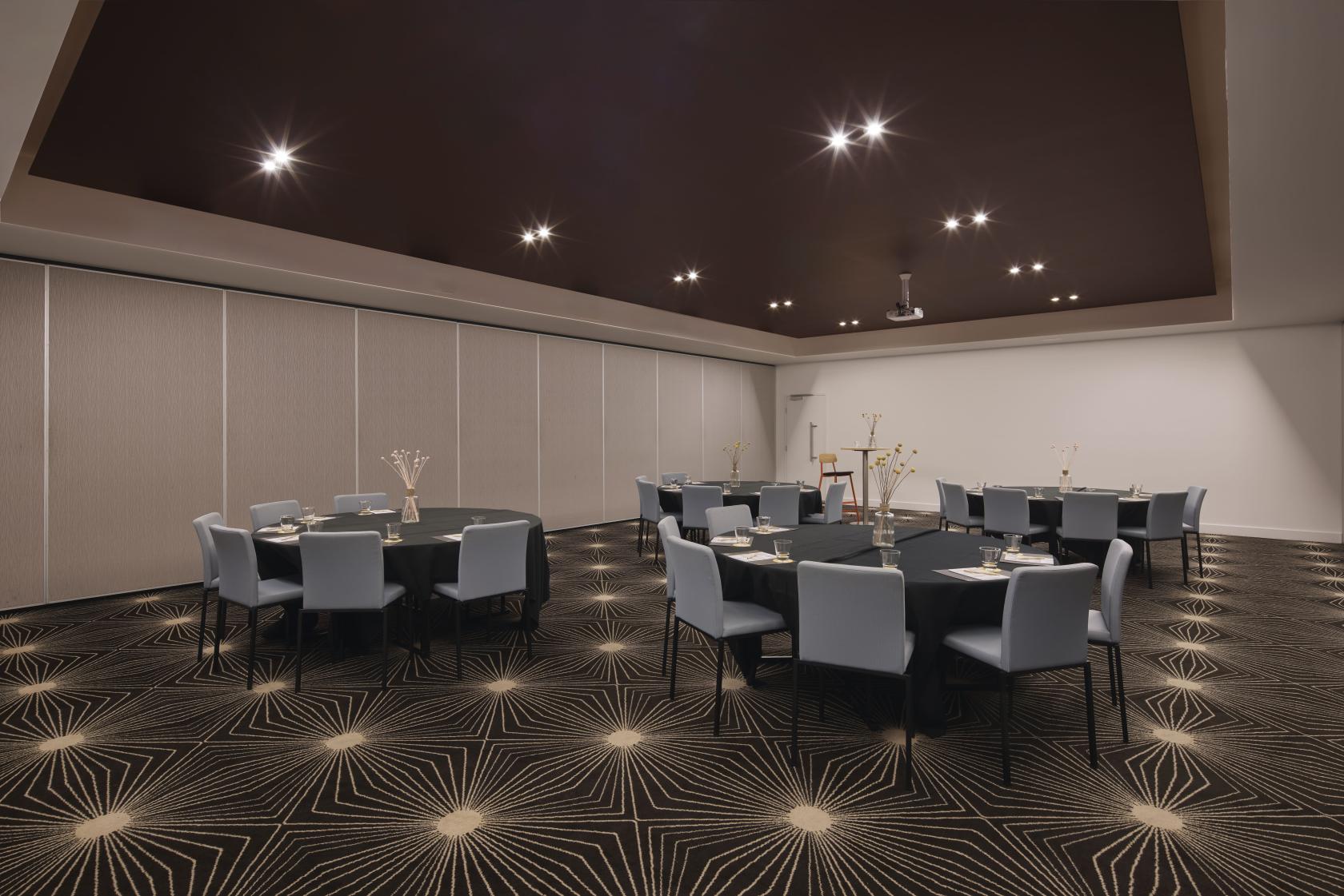 An event space with round tables, gray chairs, patterned carpet, and recessed lighting at Atura Dandenong.