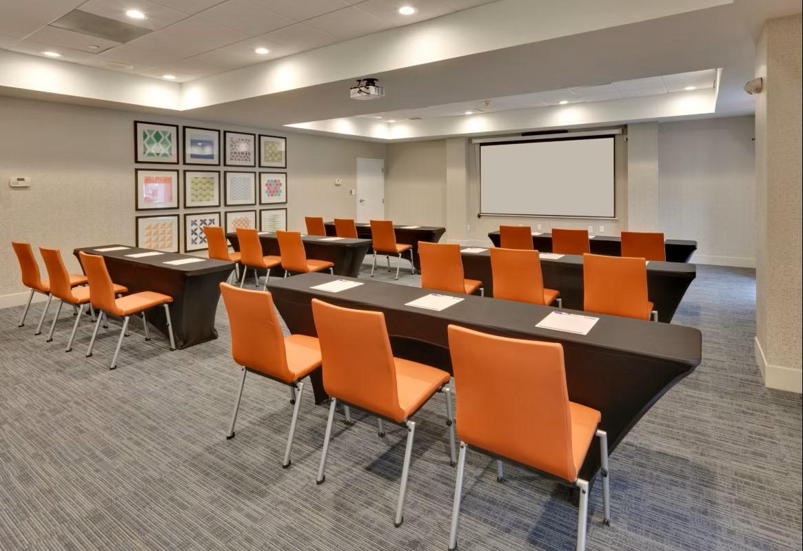 A hotel meeting room with orange chairs, black tables, a projector screen, and framed artwork at Holiday Inn Express Irving Convention Center.