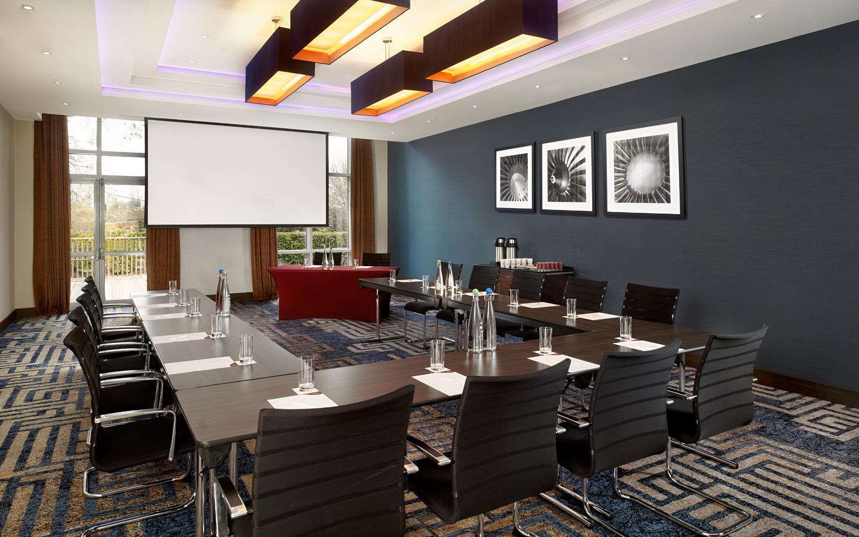 A hotel meeting room with U-shaped tables, black chairs, a projector screen, and large windows at Crowne Plaza London Gatwick.