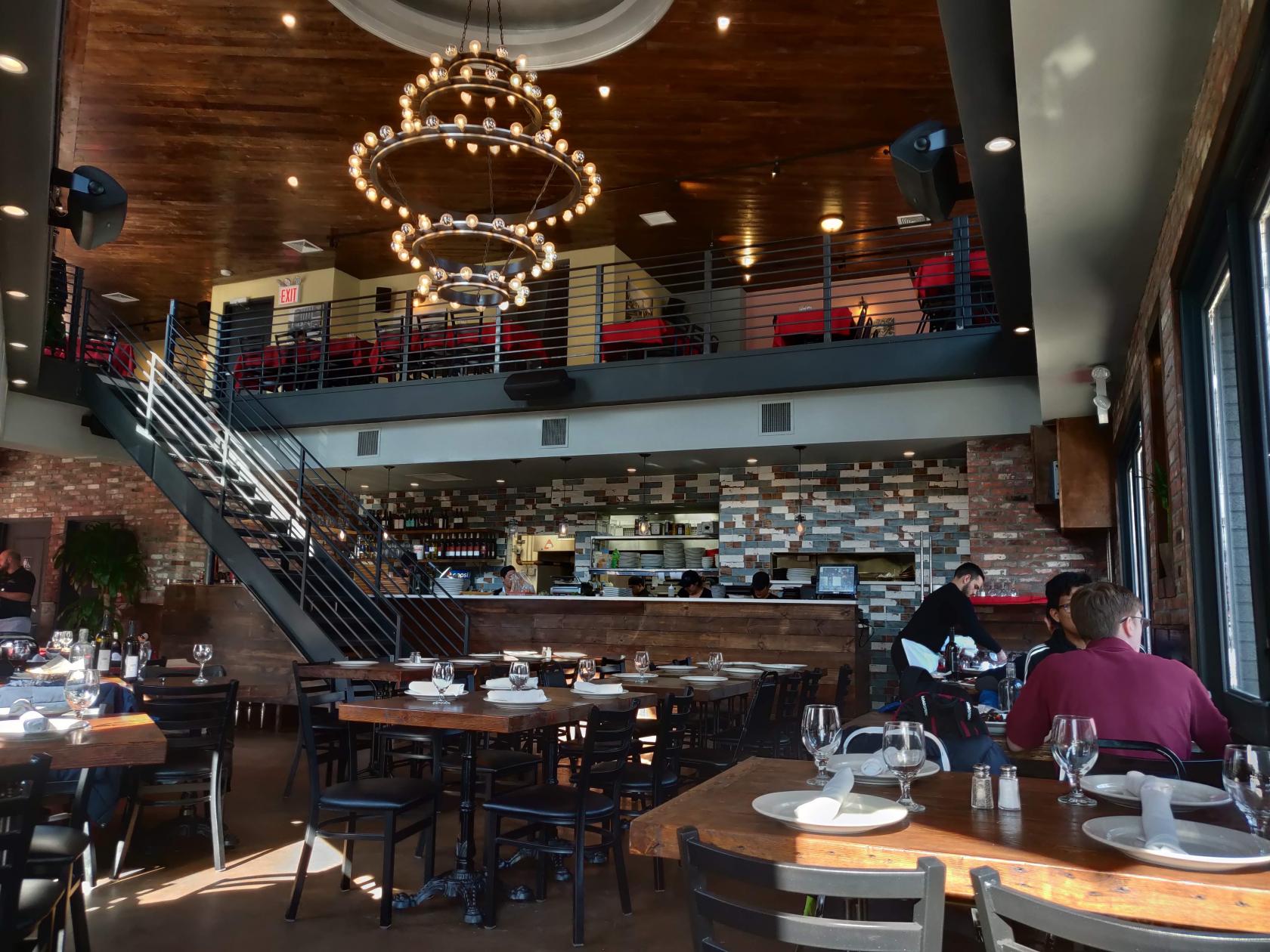 A restaurant interior with exposed brick walls, modern chandeliers, and a mezzanine level at Patrizia's of Maspeth.