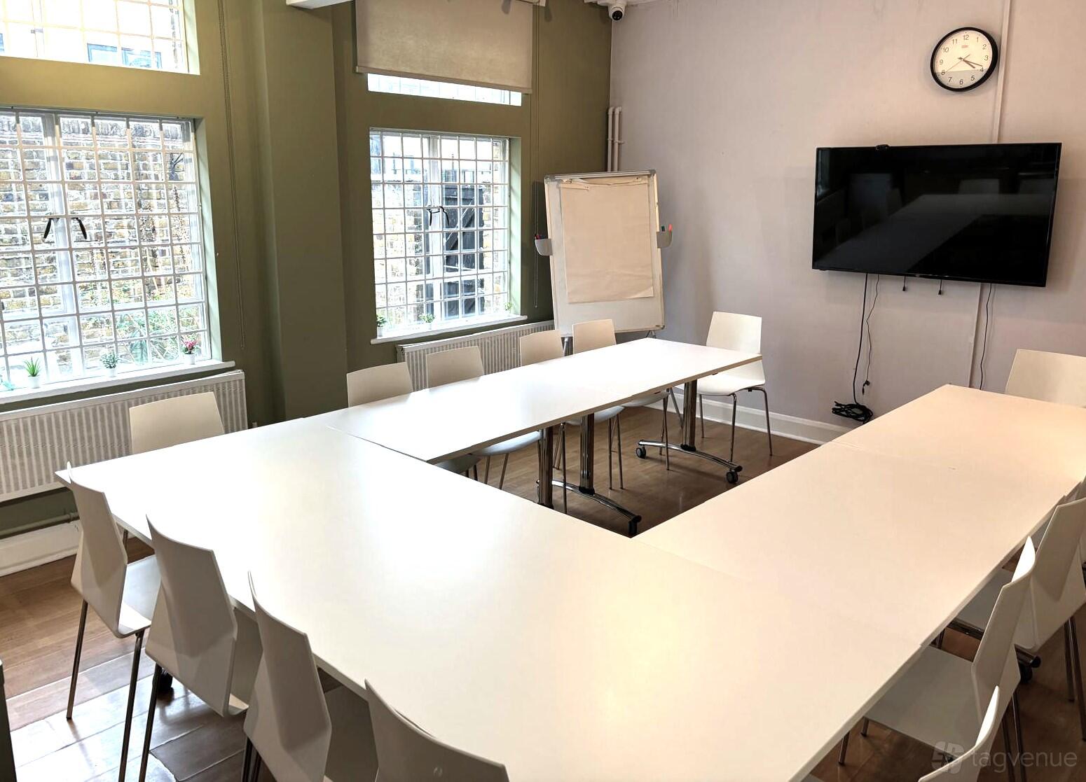 A meeting room with U-shaped white tables, white chairs, large windows, a wall clock, and a TV at Kennington Space Hire.