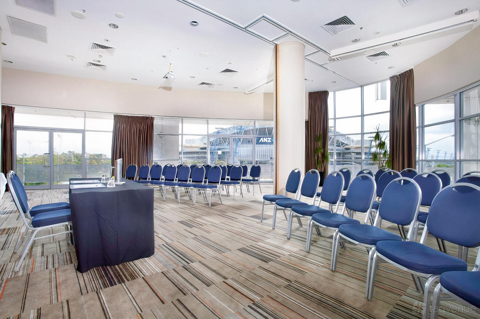 A meeting room with floor-to-ceiling windows, natural light, and rows of blue chairs at Novotel Sydney Olympic Park Hotel.