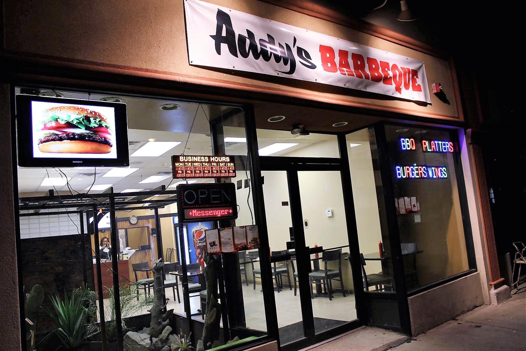 A restaurant exterior with large front windows, neon signs, and a display showing a burger at Addy's Barbeque.