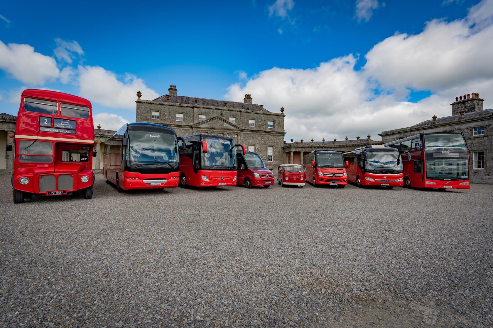 A fleet of red buses, including a vintage double-decker and modern coaches, parked at Dualway Vintage Bus.