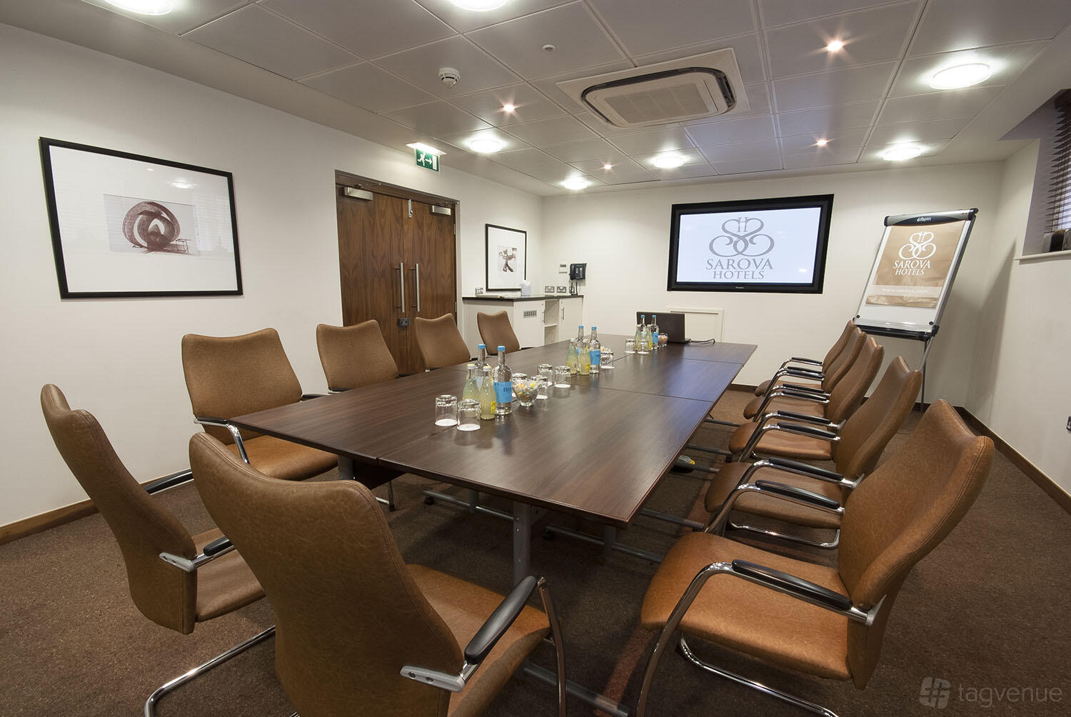 A meeting room with a long rectangular table, brown chairs, a large screen, and bottled water at Sir Christopher Wren Hotel.