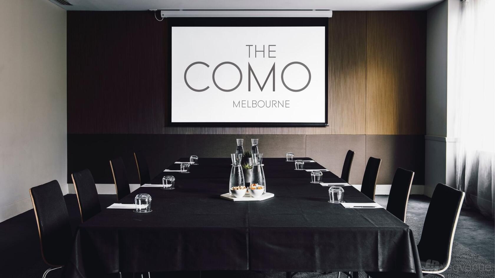 A hotel conference room with a long table, black chairs, glass water jugs, and a projector screen at The Como Melbourne.