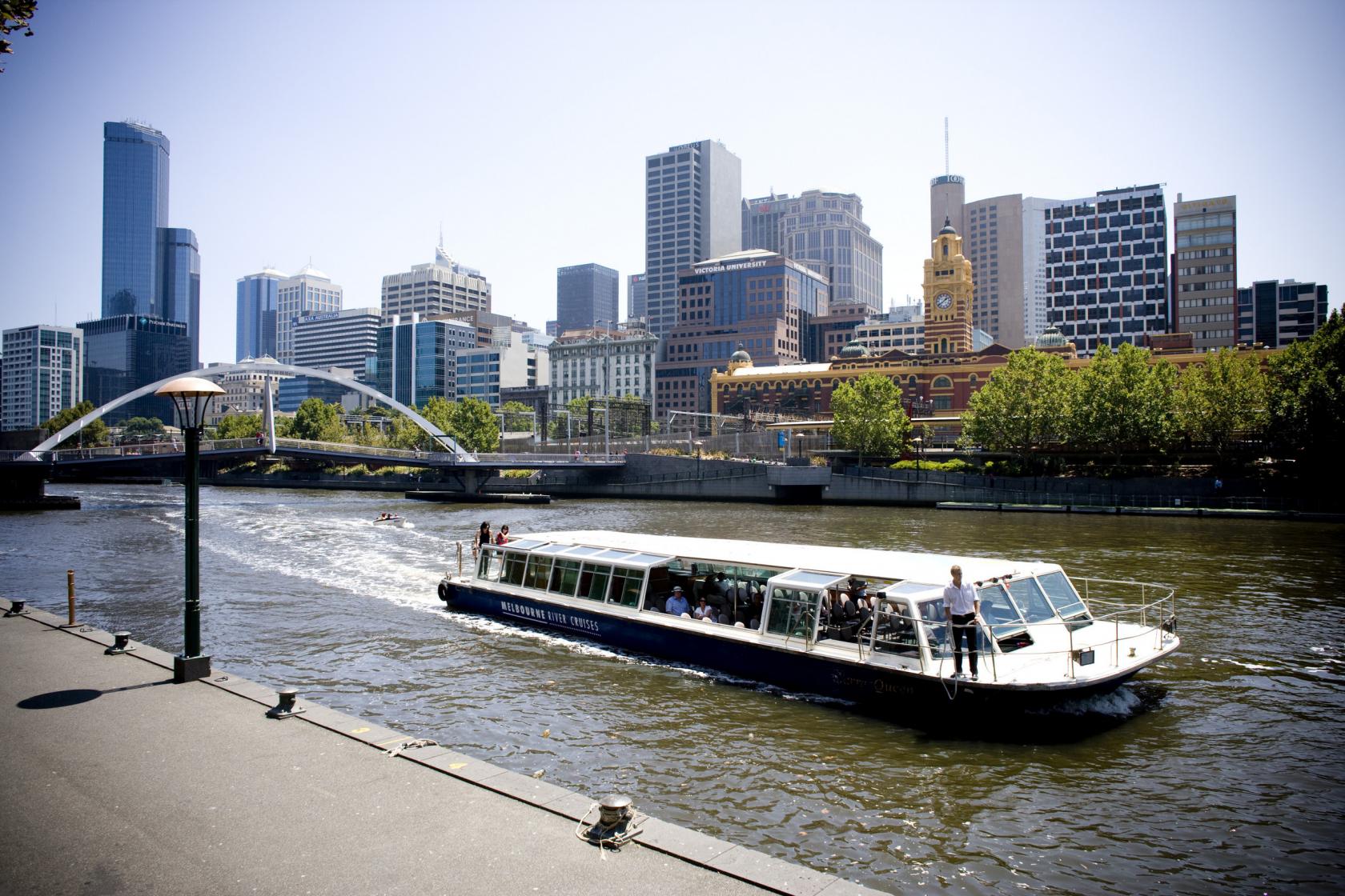 A boat with glass windows and open deck cruising on the river with city skyline views at Yarra Queen.