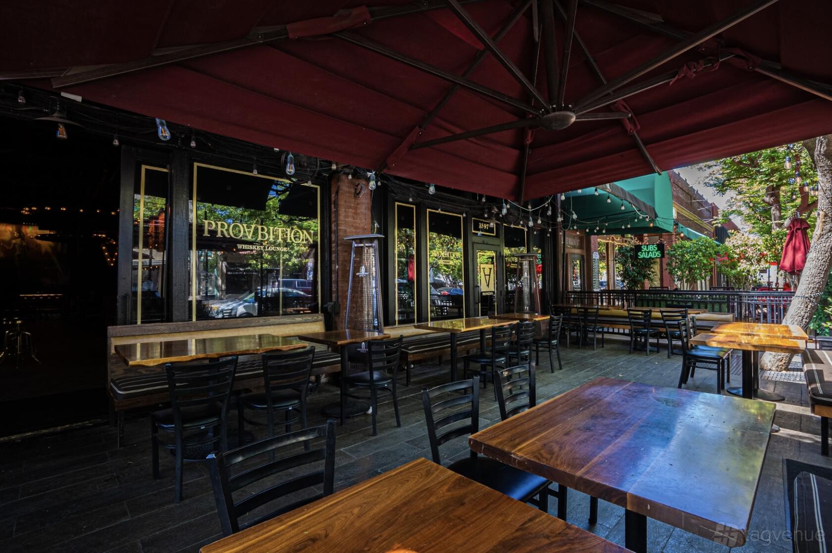 An outdoor restaurant patio with wooden tables, black chairs, and large red umbrellas at ProAbition - Riverside.