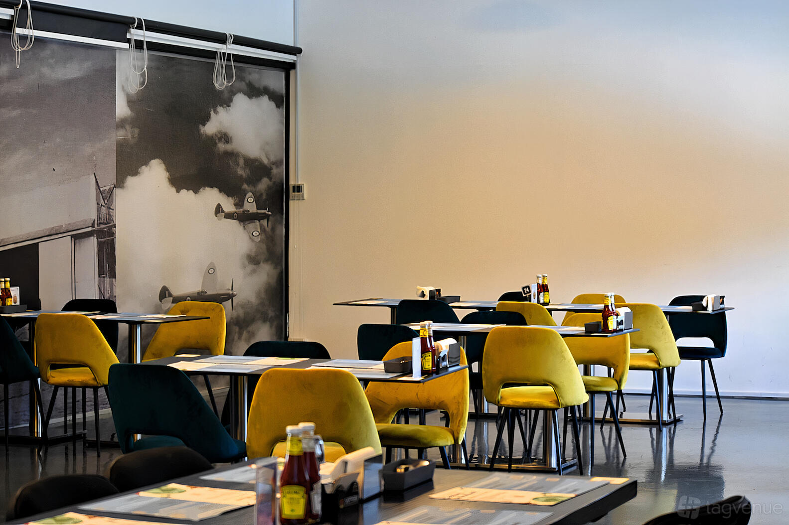 An event space with yellow and teal chairs, small dining tables, and a vintage aviation mural at WingsOverAsia.
