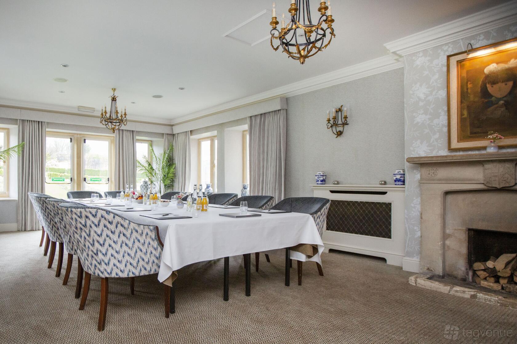 An event space with a long table, patterned chairs, chandeliers, large windows, and a stone fireplace at Lords of the Manor.