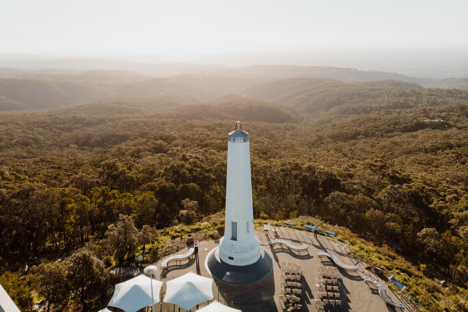 An outdoor cafe area with hillside views, white canopy umbrellas, and a tall lookout tower at Mt Lofty Summit Restaurant And Cafe.