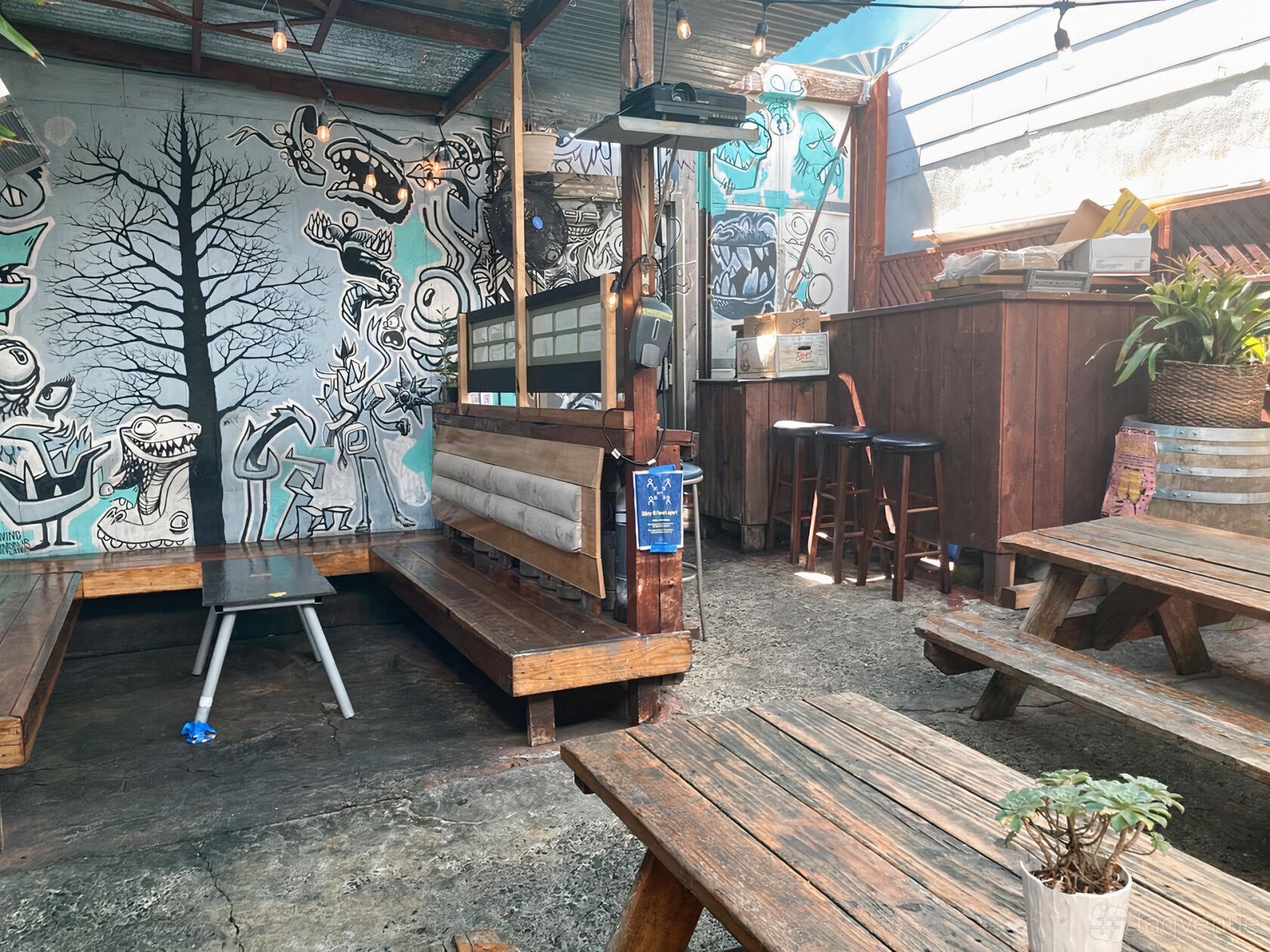 An outdoor bar area with wooden picnic tables, benches, a mural wall, and a small bar counter at The Sycamore.