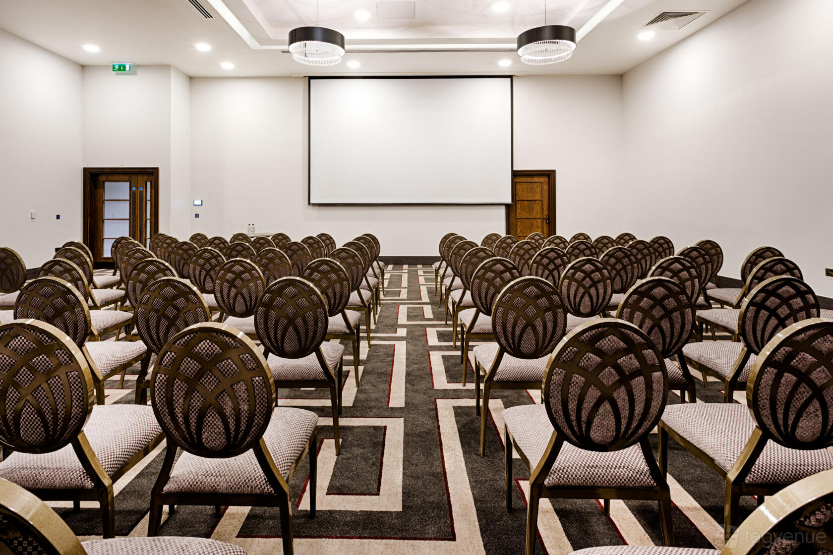 A hotel meeting room with rows of patterned chairs, geometric carpet, and a ceiling-mounted projector at Courthouse Hotel Shoreditch.