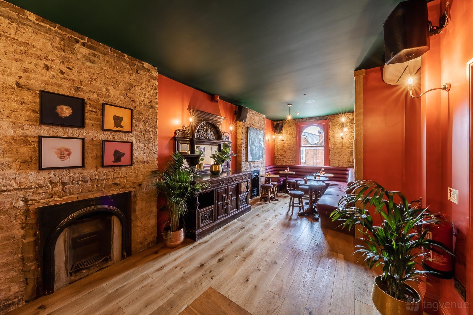 A pub room with exposed brick walls, wood floors, vintage fireplace, dark wood bar, and red banquette seating at Queen of the South.