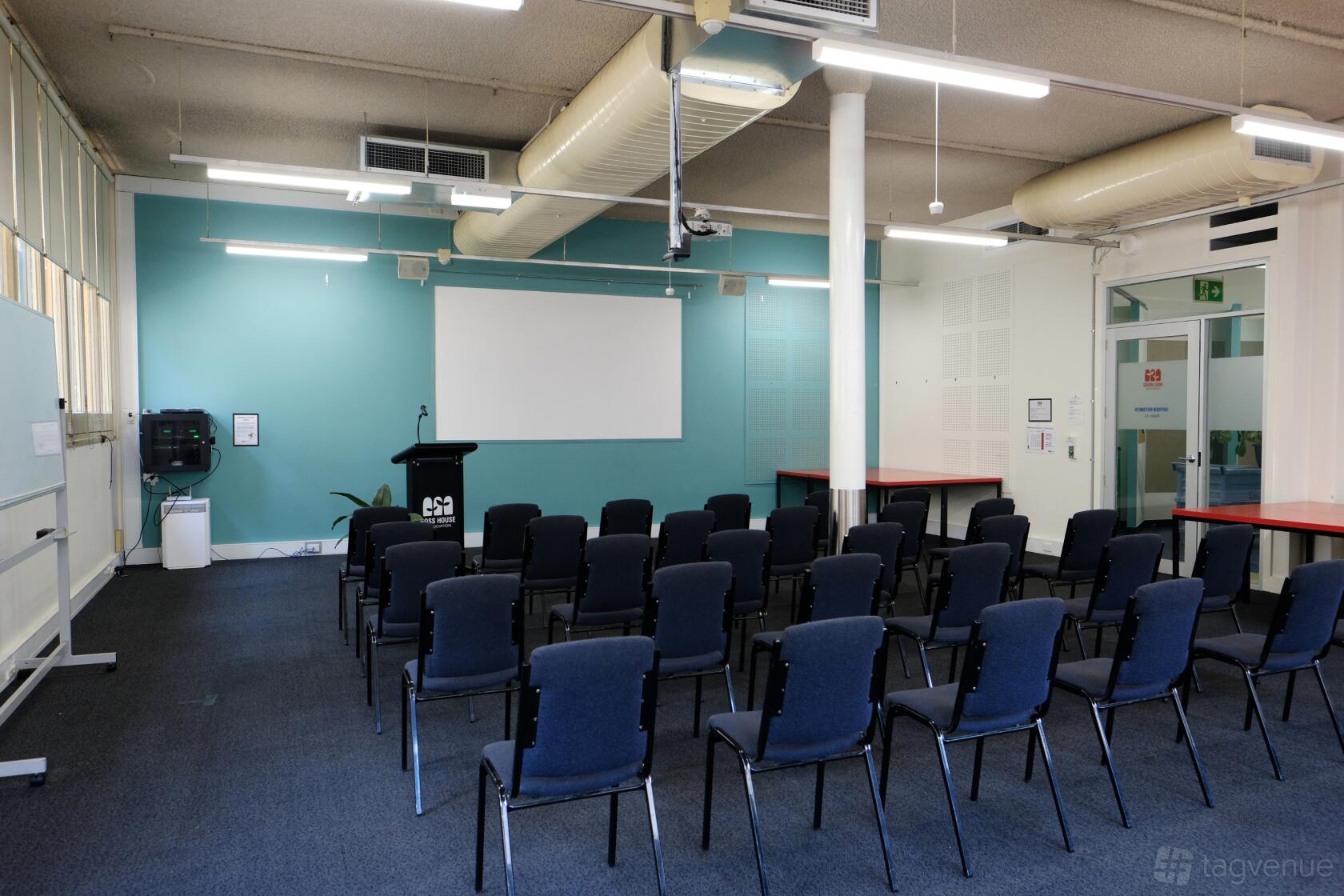 A meeting room with rows of black chairs, a podium, projector screen, and teal accent wall at Ross House.