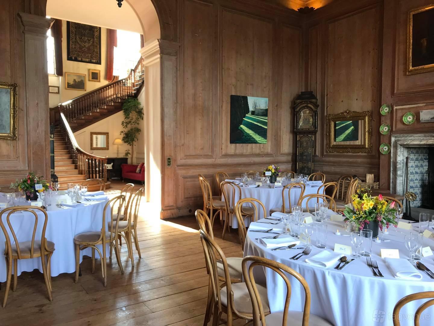 A hall with wood-paneled walls, round tables with white linens, and a grand staircase at Glemham Hall.